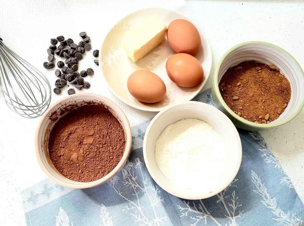 Coconut flour brownies ingredients laid out on a white and blue surface. Eggs and butter on a plate, cocoa powder, coconut flour and coconut sugar in three bowls, chocolate chips and a whisk on cloth.