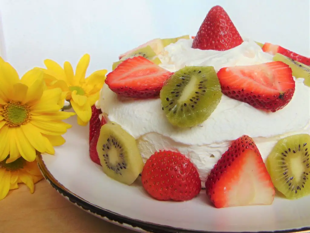 Coconut cake with whipped cream, strawberries and kiwis on a white plate and decorative yellow flowers
