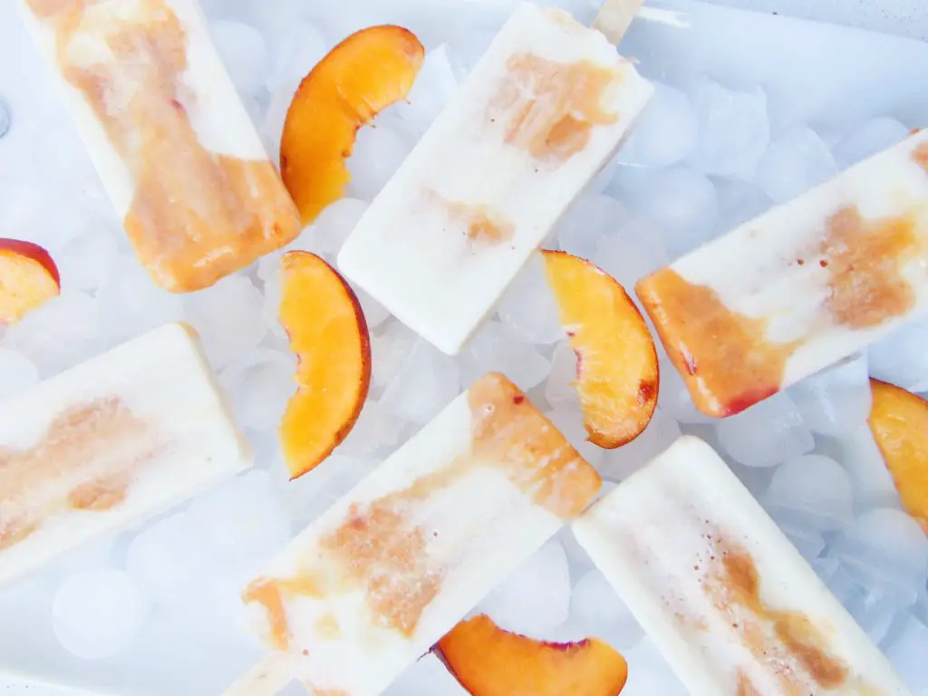 Creamy yogurt, banana and peach layered popsicles laying on ice and sliced peach as decoration