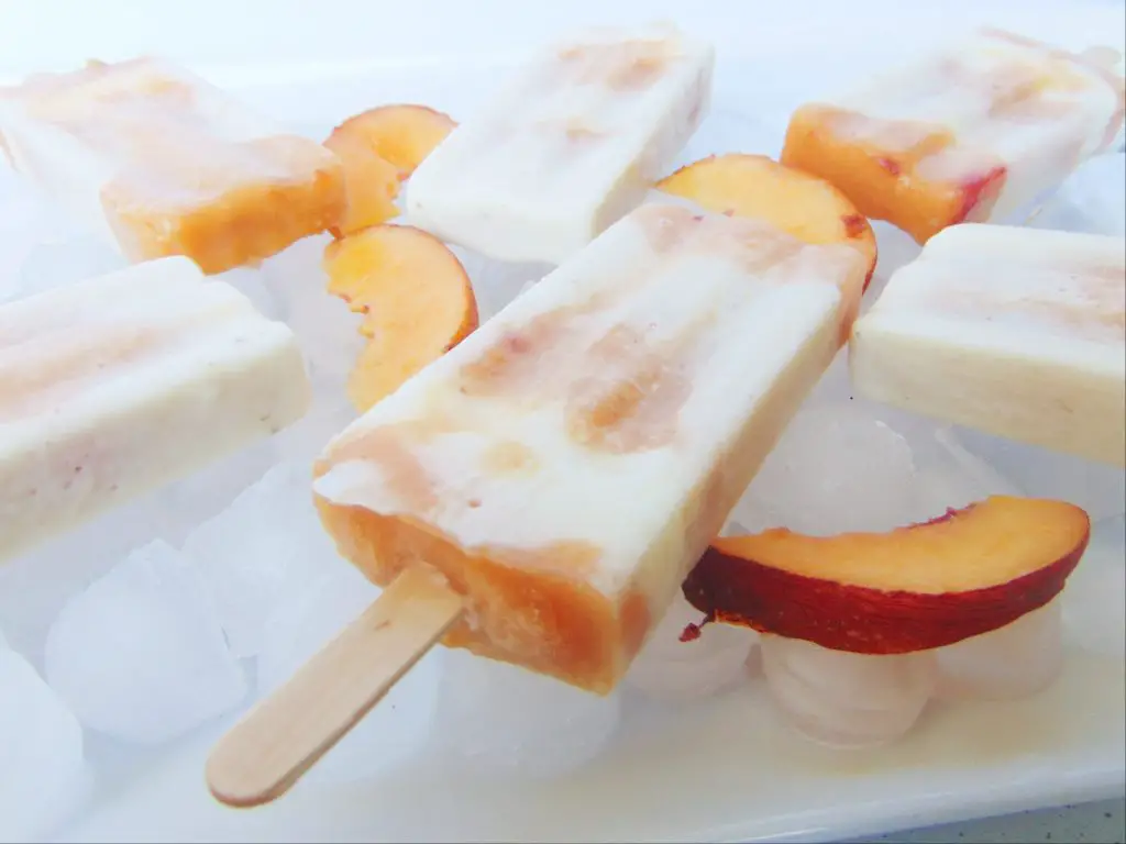 Creamy yogurt, banana and peach layered popsicles laying on ice and sliced peach as decoration