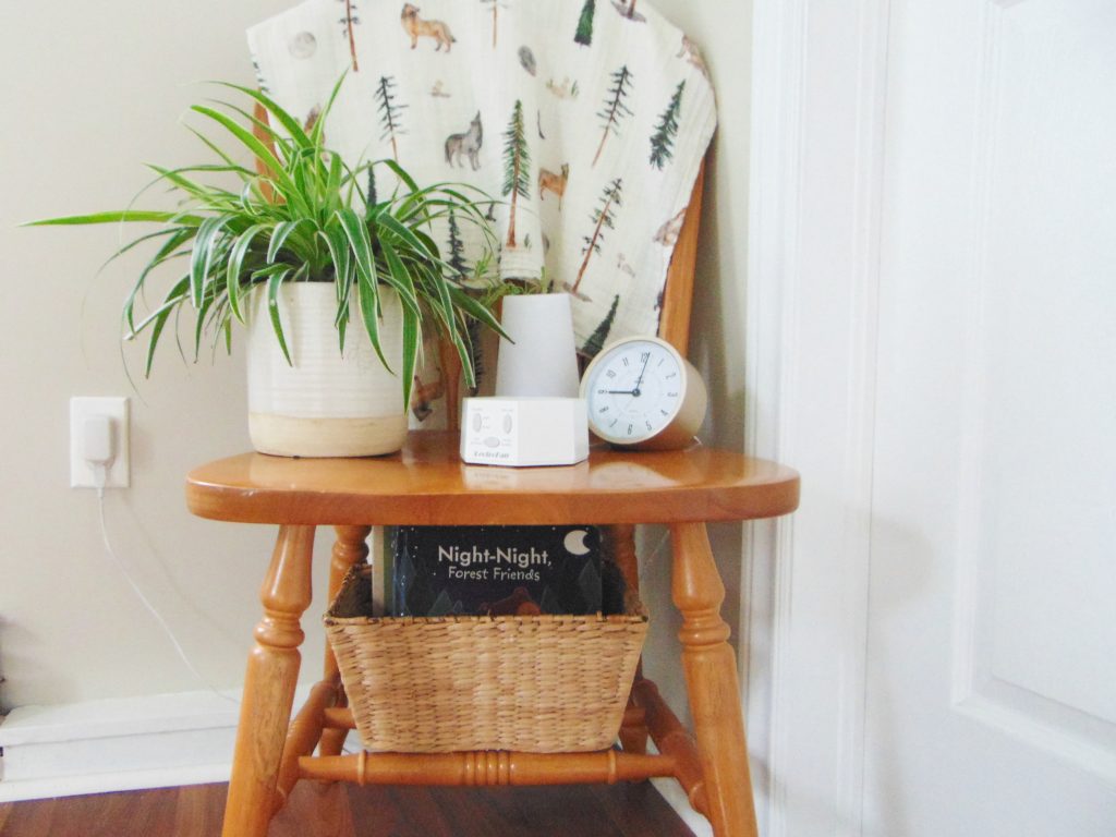 Wooden chair with forest theme blanket, plant, light, clock, sound machine, and wicker baskets full of books used as nightstand for a minimalist kid's bedroom on a budget