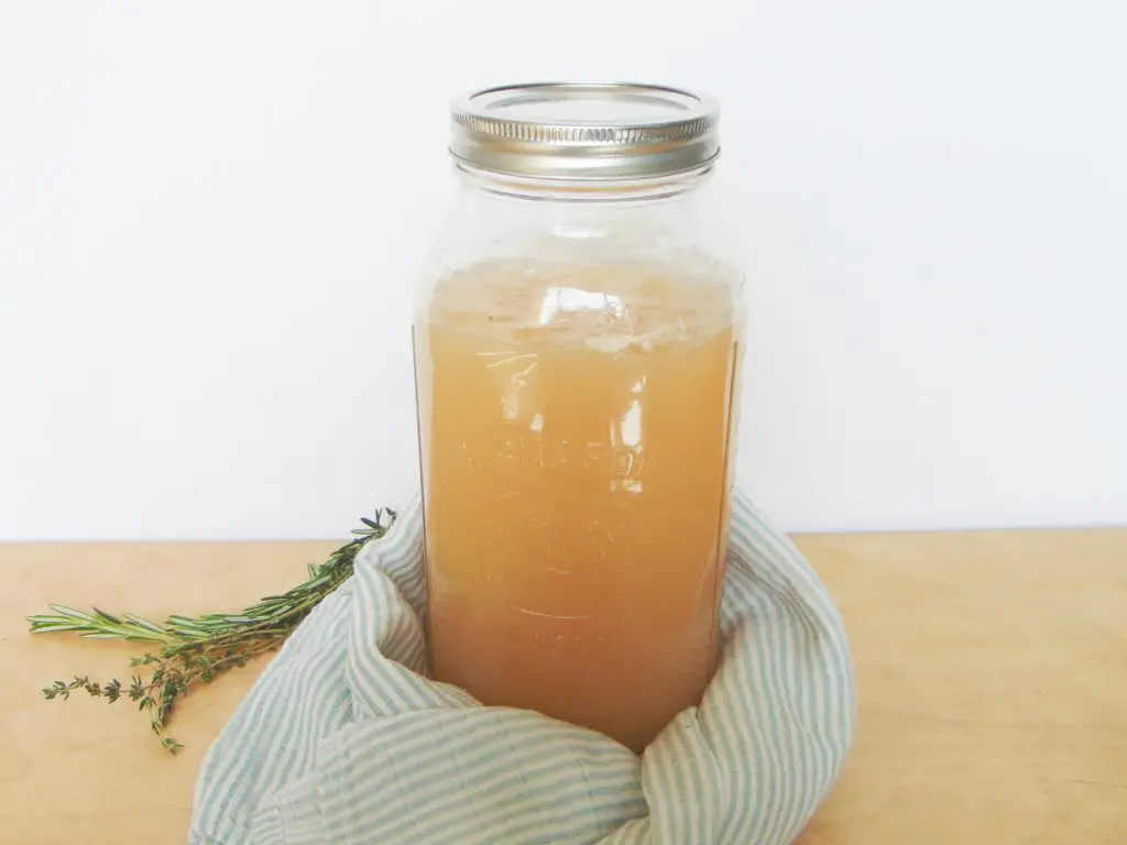 Bone broth in a mason jar on wooden surface and white background, with white and blue cloth and herbs as decoration