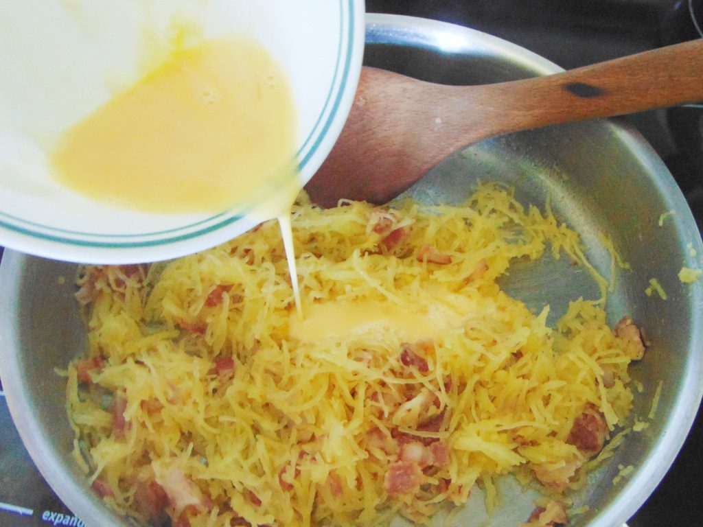 Bacon and spaghetti squash in stainless steel pan and beaten egg and milk being added to the pan