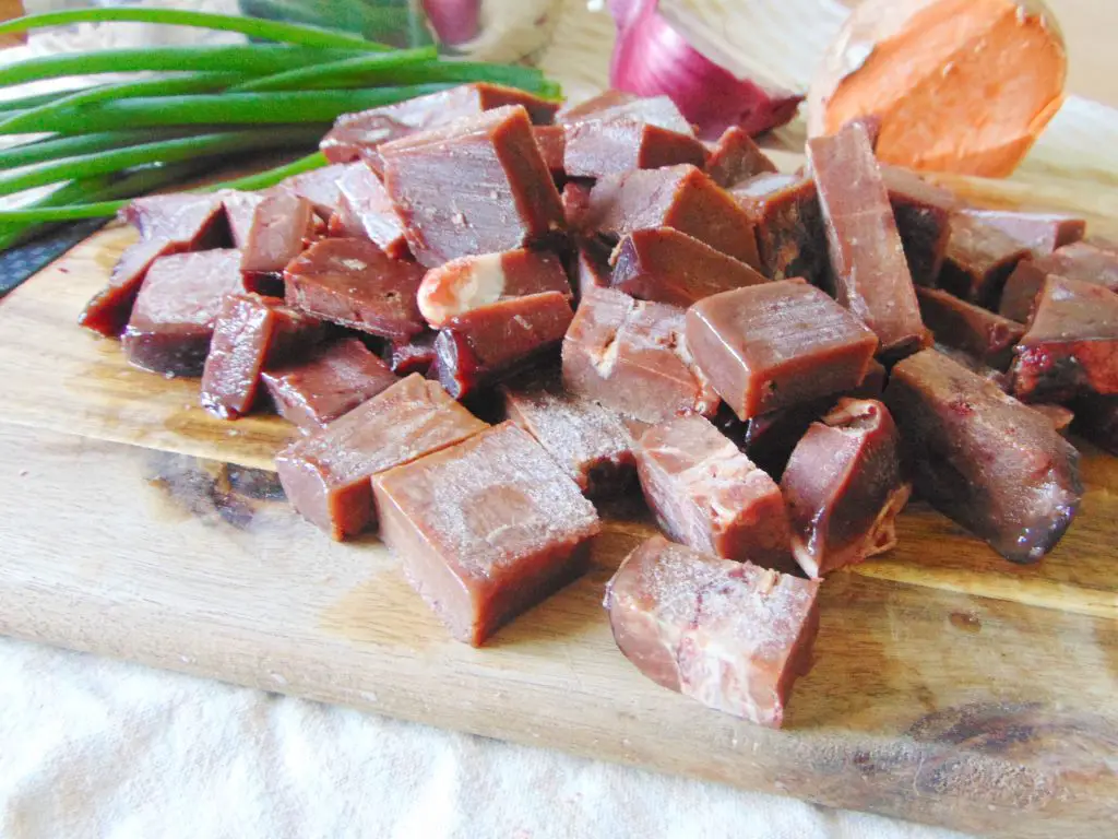 Raw beef liver chunks on wooden cutting board, with red onion, sweet potato, and green onion in background