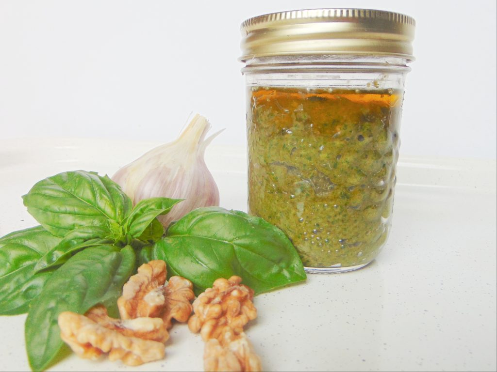 Homamade walnut pesto in small glass jar and basil, walnuts and garlic all laying on white surface