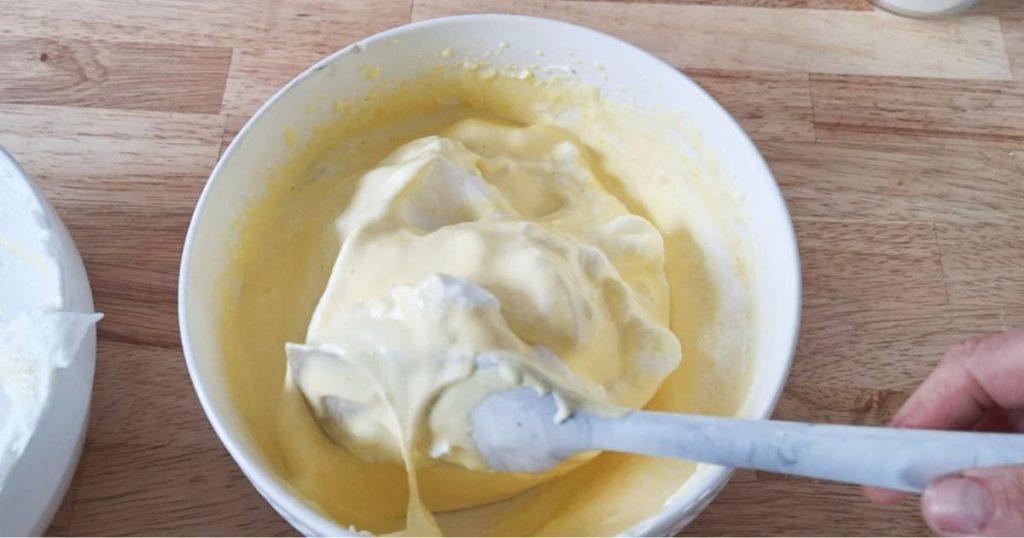 Hand gently folding soft peaks egg whites into egg yolks using a silicone spatula in a white large bowl on wooden counter