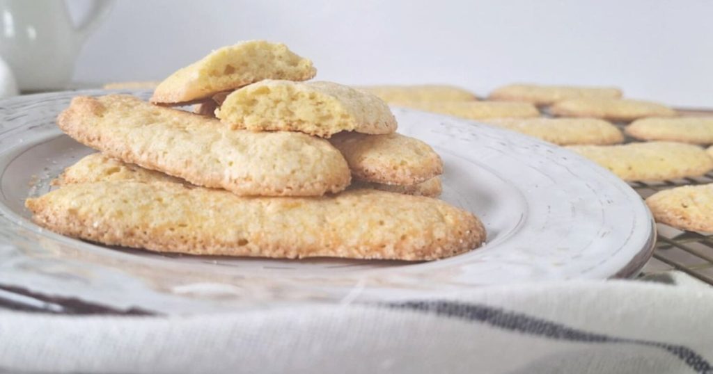 Freshly baked grain-free ladyfingers stacked on a white and brown plate with additional cookies cooling on a rack in the corner striped cloth nearby