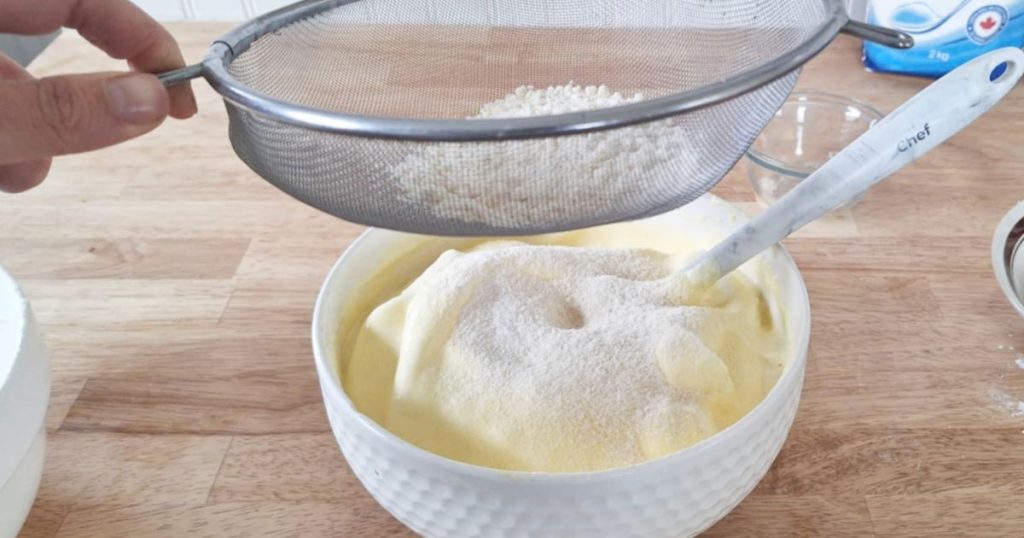Hand sifting coconut flour with a metal strainer over fluffy egg mixture in a white bowl to make grain-free ladyfingers