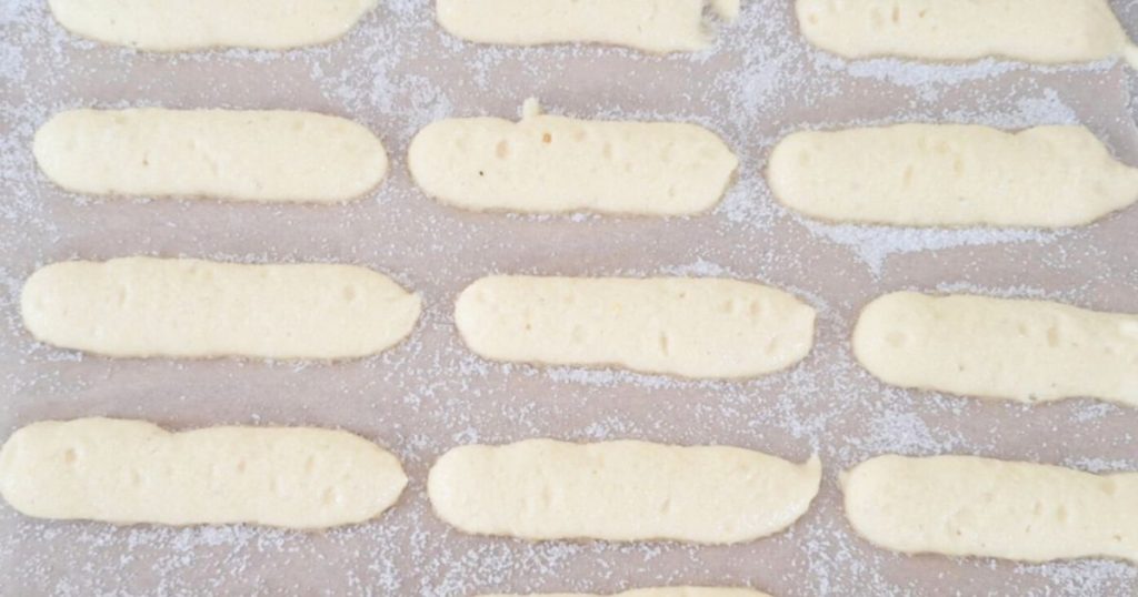 Freshly piped ladyfingers covered in sugar on baking tray lined with parchment paper ready to be baked