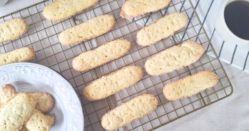 Freshly baked grain-free ladyfingers cooling on a wire rack, with a plate of additional cookies in the corner and a cup of black coffee on a striped cloth nearby