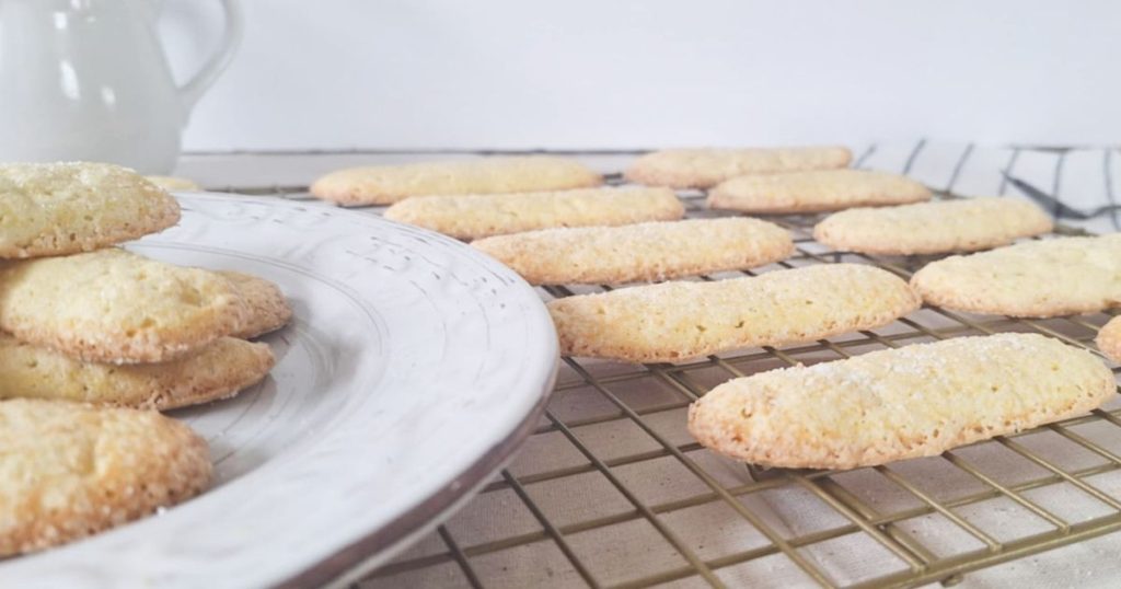 Freshly baked grain-free ladyfingers cooling on a wire rack, with a plate of additional cookies in the corner and a white pitcher in backgound