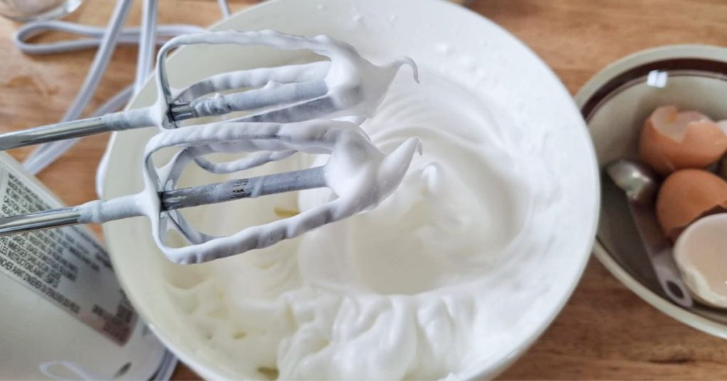 Close up of beaters with egg whites beaten to soft peaks and remaining whites in bowl and bowl with eggshells in background