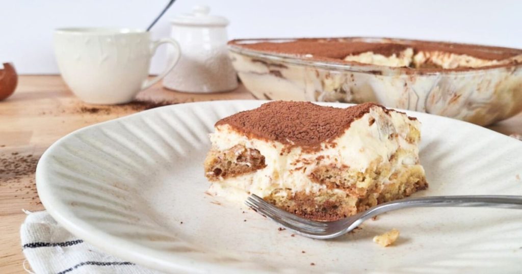 Close up of a serving of grain-free tiramisu on a white plate with remaining tiramisu in a glass dish in background and cup of coffee and sugar jar nearby on wodden counter