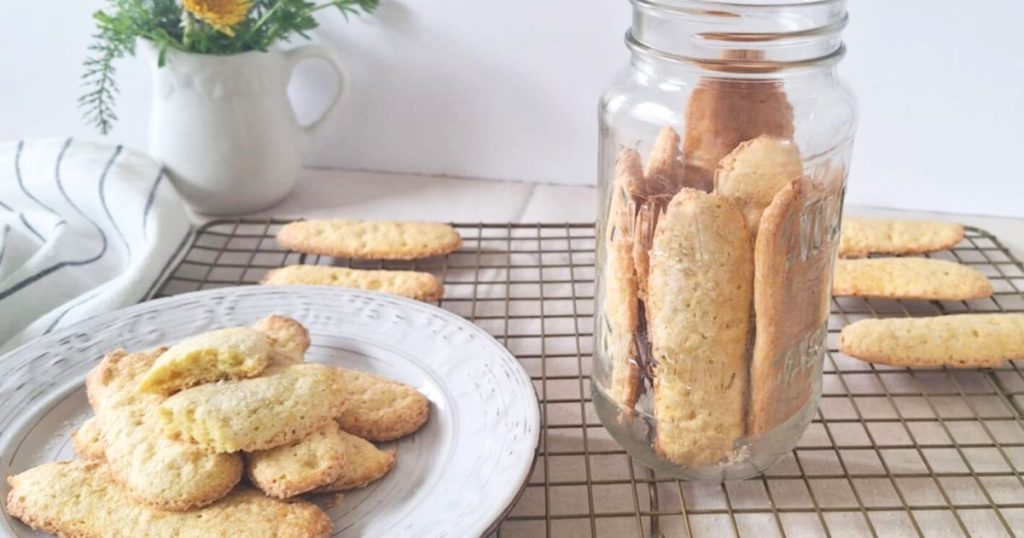 Homemade grain-free ladyfingers in a glass jar and remaining ladyfingers nearby on a plate and in background on cooling rack. Small pitcher with flowers and striped cloth in background