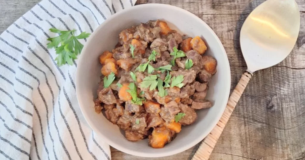 Bowl of beef heart and kidney stew garnished with fresh parsley. The stew features tender chunks of meat and carrots in a rich broth, served on a rustic wooden surface with a striped linen and a golden serving spoon.