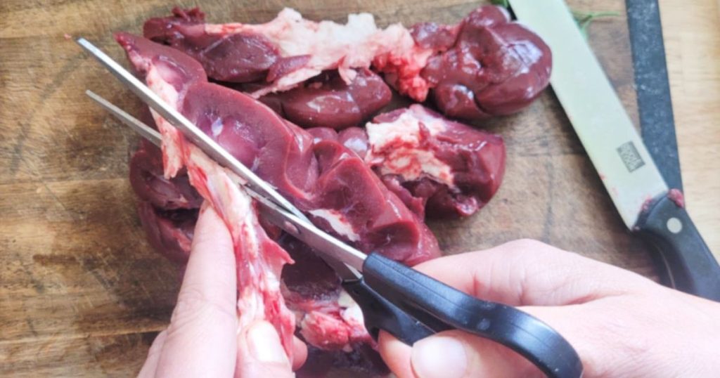 Hands trimming inner raw beef kidney fat and connective tissues with kitchen shears on a cutting board