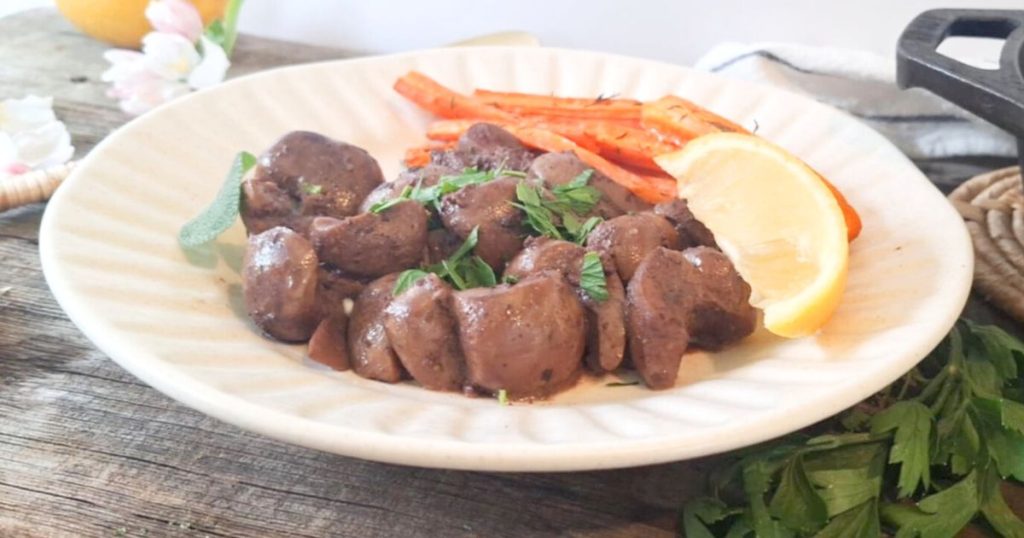 A white plate with pan-fried beef kidney pieces garnished with fresh parsley, served with roasted carrot sticks and a lemon wedge on rustic wooden table