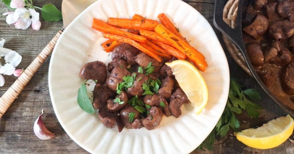 A white plate with pan-fried beef kidney pieces garnished with fresh parsley, served with roasted carrot sticks and a lemon wedge. A cast iron skillet with more cooked kidney sits to the side, on a rustic wooden table with fresh herbs, garlic, and blossoms scattered around.