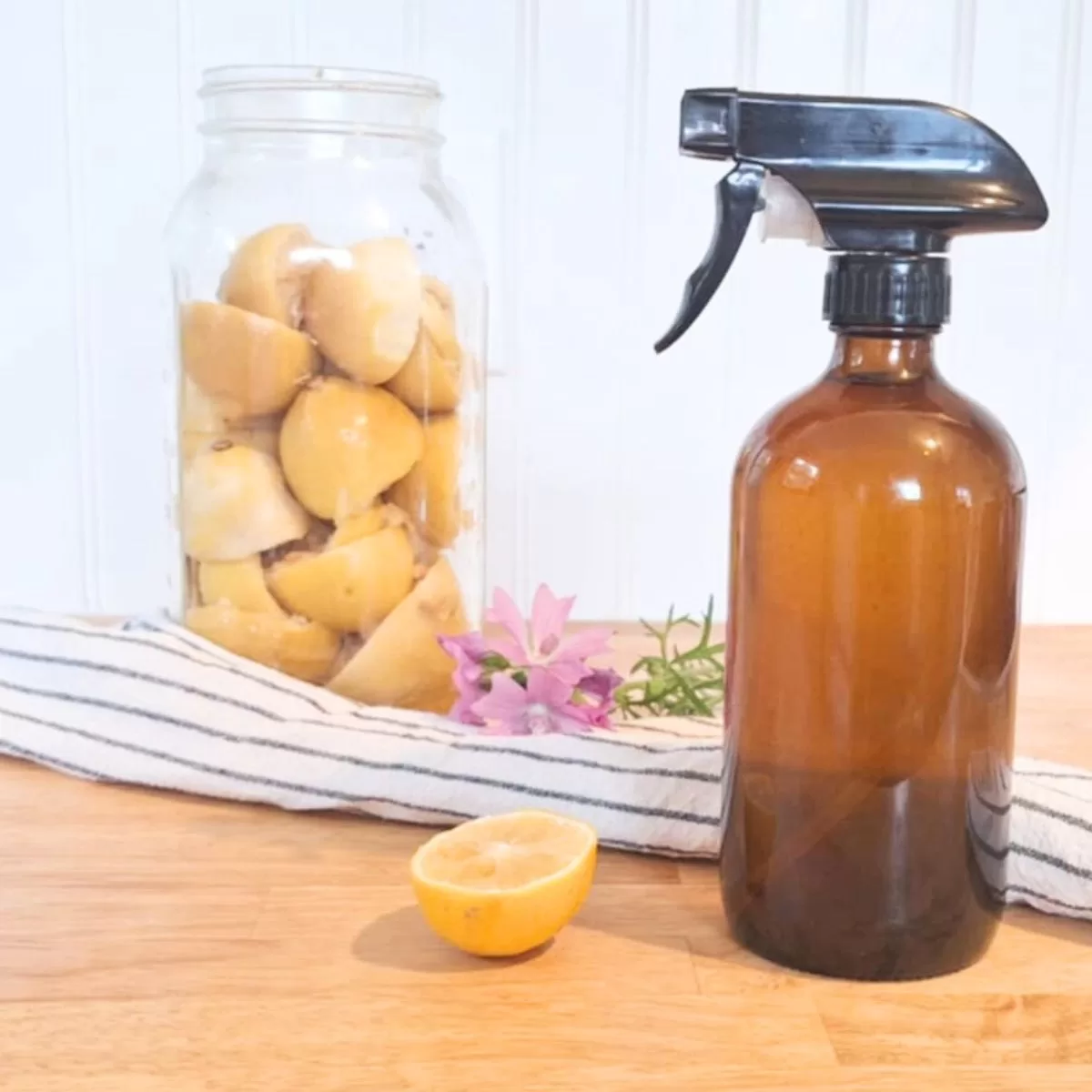 DIY lemon and vinegar cleaner in amber spray bottle with mason jar with lemon scraps, pink flowers, halved lemon and striped cloth in background